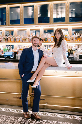 bride sits on bar while groom stands in Peacock Room at Kimpton Hotel Fontenot